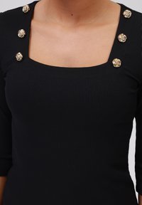 Zwarte ribbeltop met een vierkante halslijn, voorzien van goudkleurige decoratieve knopen op de schouders. Getailleerd ontwerp met driekwartmouwen.