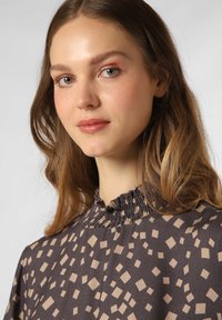 Dunkle Bluse mit Rüschenhals, verziert mit beigen geometrischen Mustern. Glatte Textur und fließendes Design. Porträtaufnahme des getragenen Kleidungsstücks.