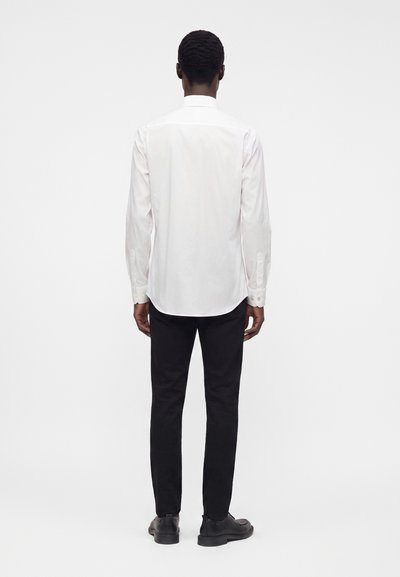 Uomo con capelli corti e ricci, di spalle, che indossa una camicia bianca a maniche lunghe, pantaloni neri e scarpe nere su sfondo semplice.