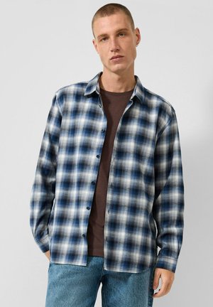 Young man wearing a blue and white checkered shirt over a brown t-shirt and blue jeans, standing against a plain light gray background.
