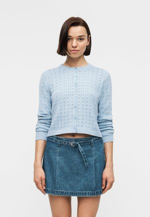 Mladá žena s krátkymi tmavými vlasmi má na sebe svetlomodrý zapínaný pletený sveter a rifľovú mini sukňu s vysokým pásom a opaskom, stojí pred jednoduchým pozadím.