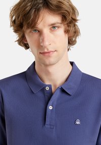 Polo de color azul marino con una tela texturizada, que presenta un cuello clásico, dos botones y un pequeño logo en el lado izquierdo del pecho.