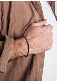 Musta kudottu rannekoru, joka on esillä ranteessa, yhdistettynä ruskeaan corduroypaitaan ja valkoiseen t-paitaan. Keskitytään tekstuuriin ja värierotukseen.