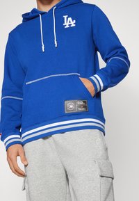 Felpa blu con lacci bianchi, logo LA sul petto, tasca a marsupio e orlo a righe. Pantaloni cargo grigi con tasche laterali.