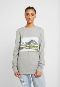 Sweatshirt cinza claro com uma ilustração frontal de montanhas e vegetação, apresentando o texto "APOIE O SEU PLANETA LOCAL." Mangas longas.