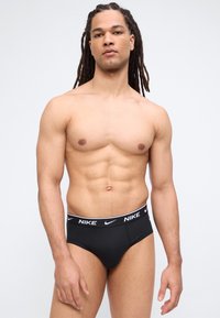 Μυώδης άνδρας με κοτσίδες που φοράει μαύρα εσώρουχα Nike, στέκεται μπροστά σε απλό φωτεινό φόντο.