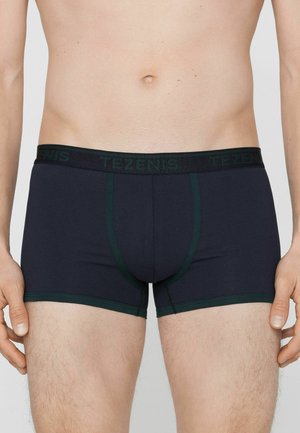 Mužský trup v tmavě modrých boxerkách se zeleným lemováním a pasem s nápisem "Tezenis" stojící na neutrálním pozadí.