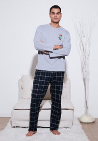 Un top de pijama gri cu mâneci lungi, având un design brodat colorat, combinat cu pantaloni din flanel cu carouri negre. Modelul stă cu brațele încrucișate.
