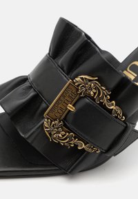 Sandal de slide em couro preto com uma tira larga, featuring uma fivela decorativa dourada e motivo floral. Textura suave e design minimalista.