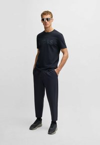 Marinblå crew neck t-shirt med präglad logo, i kombination med mörka skräddarsydda byxor och svarta sneakers. Modellen bär reflekterande solglasögon.