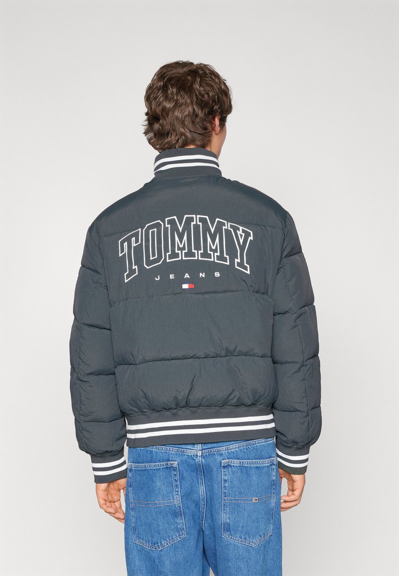 Chaqueta de abrigo gris con el texto blanco "TOMMY JEANS" en la parte posterior. Presenta puños y dobladillo acanalados y a rayas. Textura suave y acolchada.