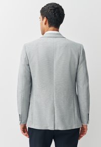 Homme aux cheveux courts et bouclés, portant un blazer gris clair et un pantalon sombre, debout dos à un fond blanc uni.