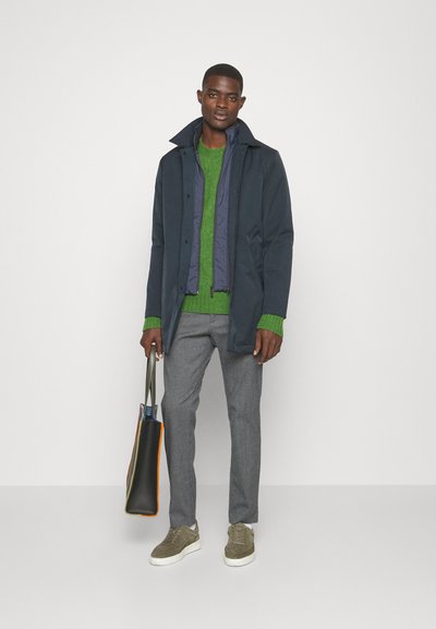 Mørkeblå frakke med høj krave over en grøn sweater, grå bukser, olivenfarvede sneakers og en sort tote bag med farvede detaljer.
