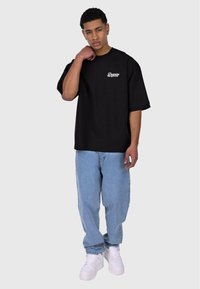 Schwarzes übergroßes T-Shirt mit kleinem weißen Logo auf der Brust, kombiniert mit hellblauen Jeans und weißen Sneakern. Einfaches, lässiges Design.