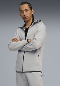 Lys grå zip-up jakke med en blød tekstur, sorte accenter og en hætte. Har sidelommer og en tætsiddende design med ribbede manchetter.