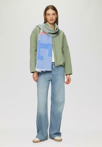 Blouson aviateur vert, T-shirt blanc, jean large bleu clair et écharpe multicolore avec des motifs géométriques et des bords frangés.