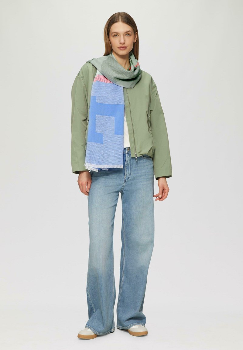 Blouson aviateur vert, T-shirt blanc, jean large bleu clair et écharpe multicolore avec des motifs géométriques et des bords frangés.