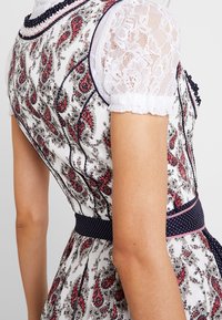 Robe blanche avec un motif paisley rouge et noir, ceinture à pois marine, et manches bouffantes en dentelle blanche transparente sur le bras et l'épaule d'une personne.