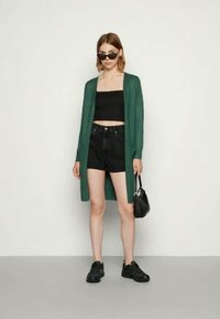 Young woman wearing black crop top, black shorts, green long cardigan, black sneakers, sunglasses, and holding small black handbag.