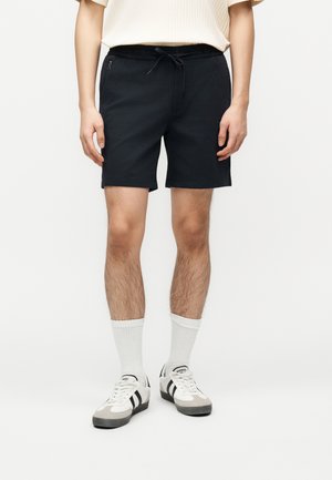Mann trägt schwarze Shorts mit Kordelzug, weiße Socken und weiße Sneaker mit schwarzen Streifen, steht vor einem schlichten Hintergrund.