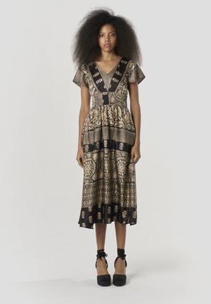 Vestido maxi con tela de patrón negro y dorado, escote en V, mangas cortas con vuelo, cintura ceñida, falda fluida y zapatos con pulsera en el tobillo.