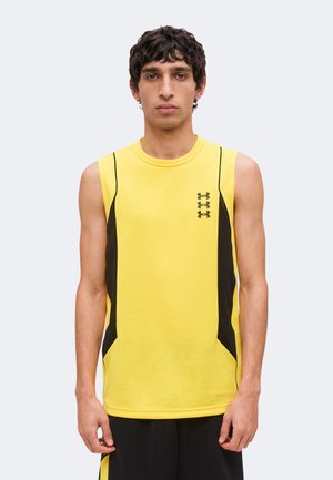 Ung mand med krøllet mørkt hår iført en gul ærmeløs sportstrøje med sorte sidepaneler og sorte shorts står med fronten mod kameraet.