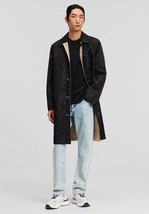 Abrigo negro impermeable con un patrón texturizado, forro de color claro y botones, combinado con jeans de color azul claro y zapatillas blancas.