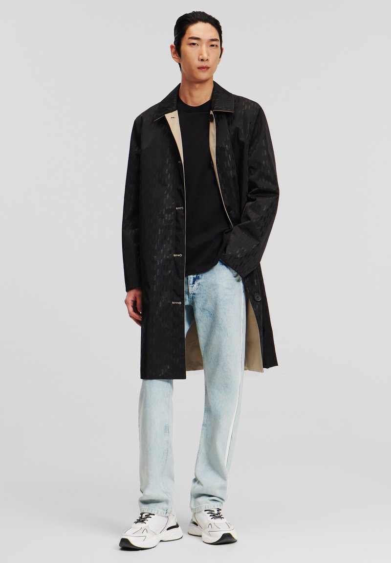 Abrigo negro impermeable con un patrón texturizado, forro de color claro y botones, combinado con jeans de color azul claro y zapatillas blancas.