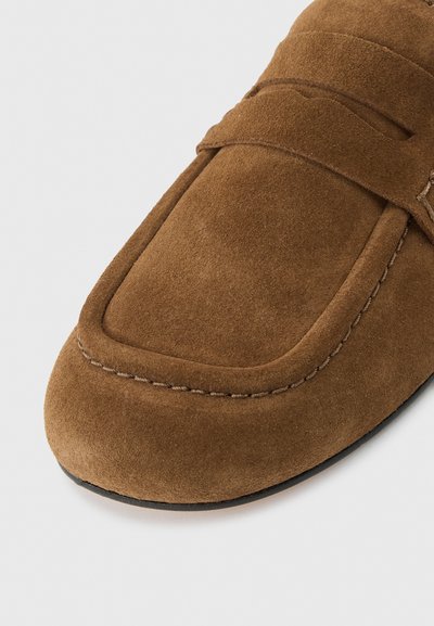 Καφέ μοκασίνι από σουέντ με ραμμένες λεπτομέρειες και λουράκι πάνω από το πάνω μέρος, εμφανιζόμενο σε ανοιχτό γκρι φόντο.