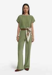 Olivgrön jumpsuit med lös passform, korta ärmar och vida ben, accentuerad med ett brunt bälte och en mönstrad scarf knuten i midjan.