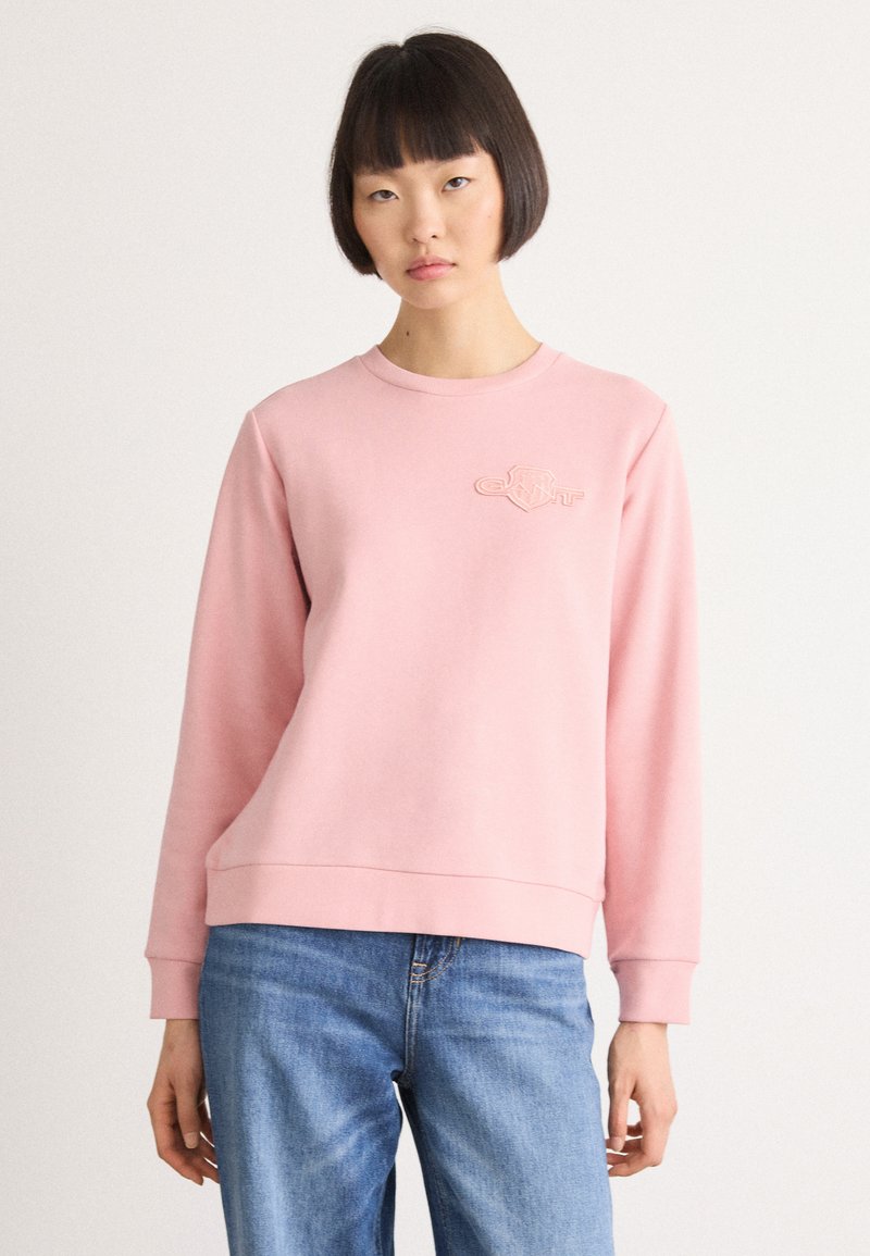 Vrouw met kort zwart haar, gekleed in een lichtroze sweatshirt met een klein logo op de borst en blauwe jeans, tegen een effen achtergrond.