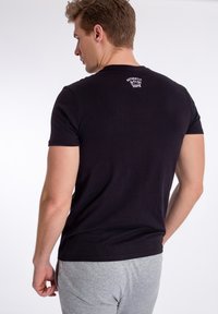 Sort kortærmet T-shirt lavet af bomuld, med rund hals og hvid tekstlogo på den øverste del af ryggen. Båret med grå bukser.