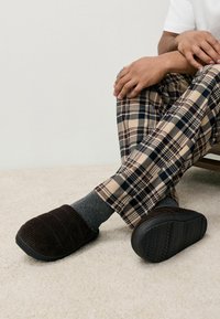 Hnedé kordurové papuče s textúrovaným povrchom, čierne gumové podrážky, nosené so sivými ponožkami a béžovými kockovanými nohavicami na svetlom koberci.