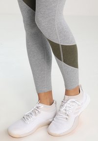Leggings sportivi grigi con dettagli geometrici verde oliva, abbinati a sneakers bianche in mesh, con suola testurizzata e lacci.