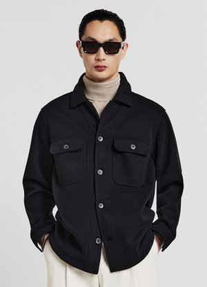 Giacca camicia in misto lana nera con due tasche sul petto e chiusura con bottoni. Indossata sopra un dolcevita beige. Modello con occhiali da sole scuri.