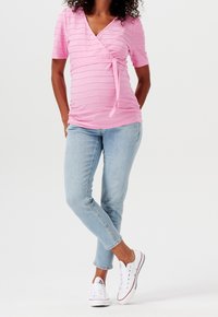 Růžový krátký top s dlouhým rukávem a horizontálními texturálními detaily, kombinovaný se světle modrými džínami a bílými teniskami. Top má zavazování v pase.