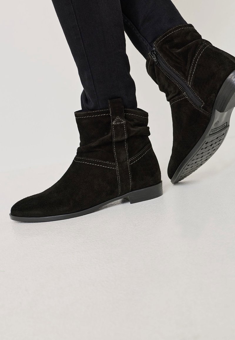 Bottines en daim noir avec fermeture éclair latérale, détails de couture et petit talon plat, portées avec un pantalon noir ajusté.