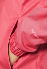 Chaqueta impermeable rosa con puños elásticos y un logo visible "DRYVENT". El material tiene una textura suave y es ligero.