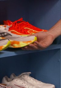 Jooksujalatsid oranžides toonides, kollaste aktsentidega ja tekstureeritud valge ja oranži tallaga, mida hoiab käsi sinisel riiulil.