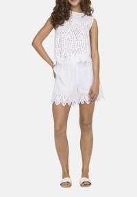 Weißes ärmelloses Oberteil mit Lochstickerei und gewellten Kanten, kombiniert mit passenden weißen Shorts, die ein ähnliches gewelltes Design aufweisen.
