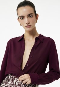Burgundy button-up shirt with a collar and deep v-neck. Features long sleeves and contrasting buttons, paired with a patterned skirt.
