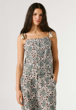 Junge Frau mit langen dunklen Haaren trägt ärmelloses, gemustertes Oberteil und passende Hose mit Seitentaschen, steht vor einfachem Hintergrund.