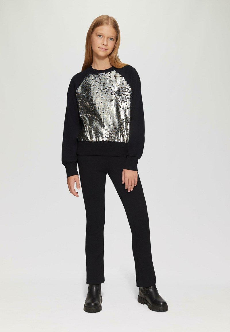Schwarzer Sweatshirt mit einer silbernen Pailletten-Front, langen Ärmeln und einem taillierten Saum. Kombiniert mit schwarzen, engen Hosen und schwarzen Stiefeletten.