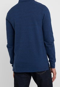 Person som bär en långärmad, mörkblå stickad skjorta och mörka jeans, stående med ryggen mot kameran.