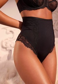 Zwarte high-waisted slips met kantafwerking, mesh-details en een naadloos ontwerp, die de gladde textuur en elegante accenten benadrukken.