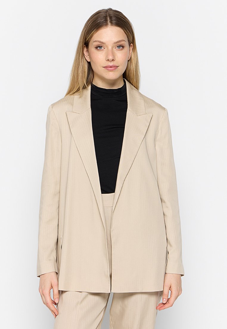 Beige maßgeschneiderter Blazer mit scharfen Revers, vertikalen Streifen und einer lockeren Passform. Über einem schwarzen Rollkragenpullover getragen, mit Seitentaschen.