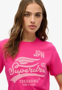 T-shirt en coton rose avec un design graphique blanc affichant "Superdry," "Yokohama," et "JPN #01." Manches courtes et style col rond.