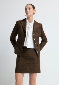 Hnědý tweedový blejzr se zlatými knoflíky a přiléhavým střihem, doplněný bílou blůzou s mašlí. Sukně je z materiálu odpovídajícího blejzru.