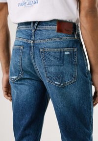Jeans de mezclilla azul con un acabado desgastado, que presentan dos bolsillos traseros y una etiqueta de cuero. Un bolsillo tiene un patrón de la bandera del Reino Unido.
