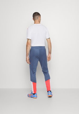 Man standing facing a wall wearing white short-sleeve shirt, blue track pants with red and white lower panels, and blue sports cleats.